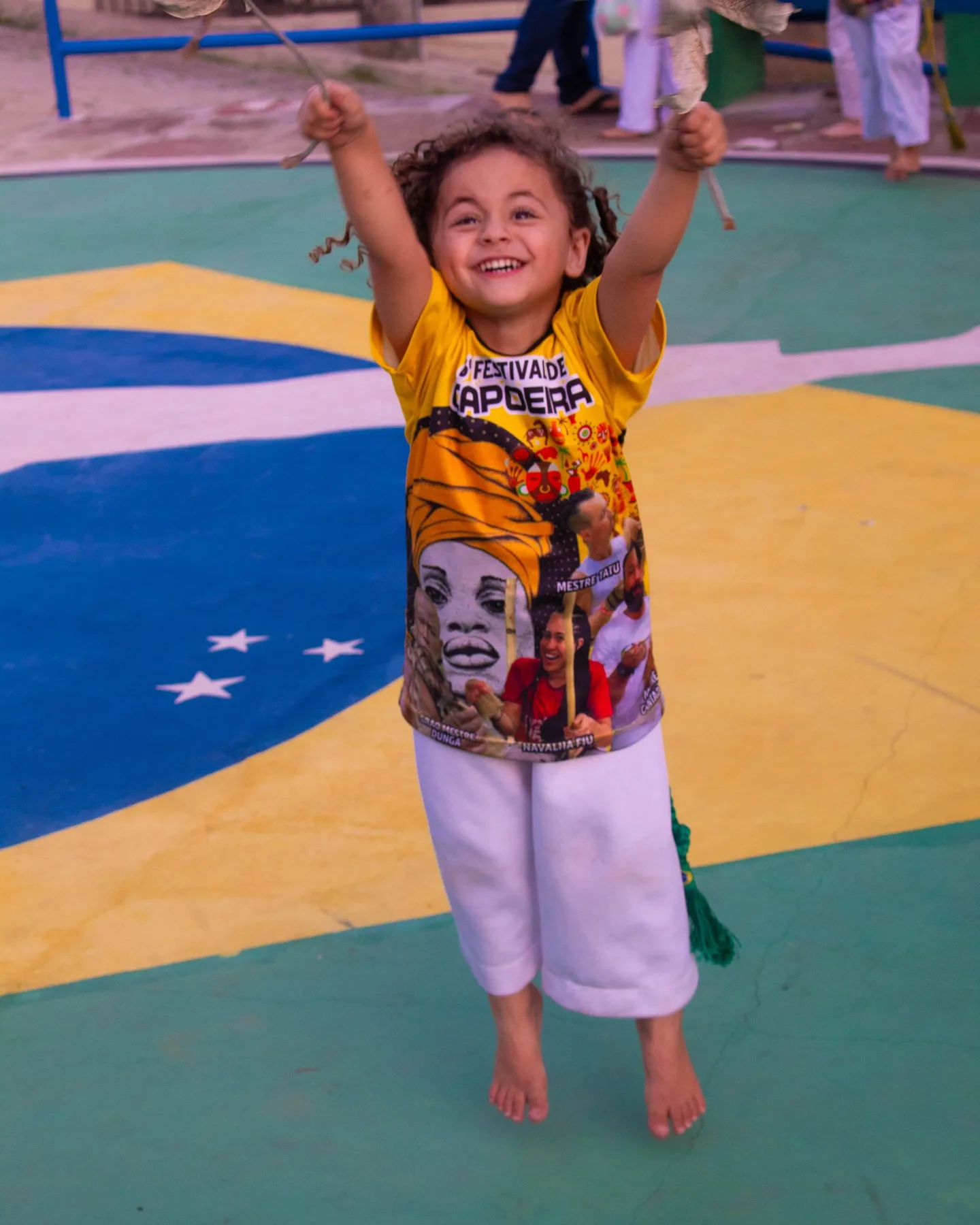 Criança de camiseta amarela do Festival de Capoeira pula com os braços erguidos, sorrindo, sobre um piso pintado com a bandeira do Brasil.
