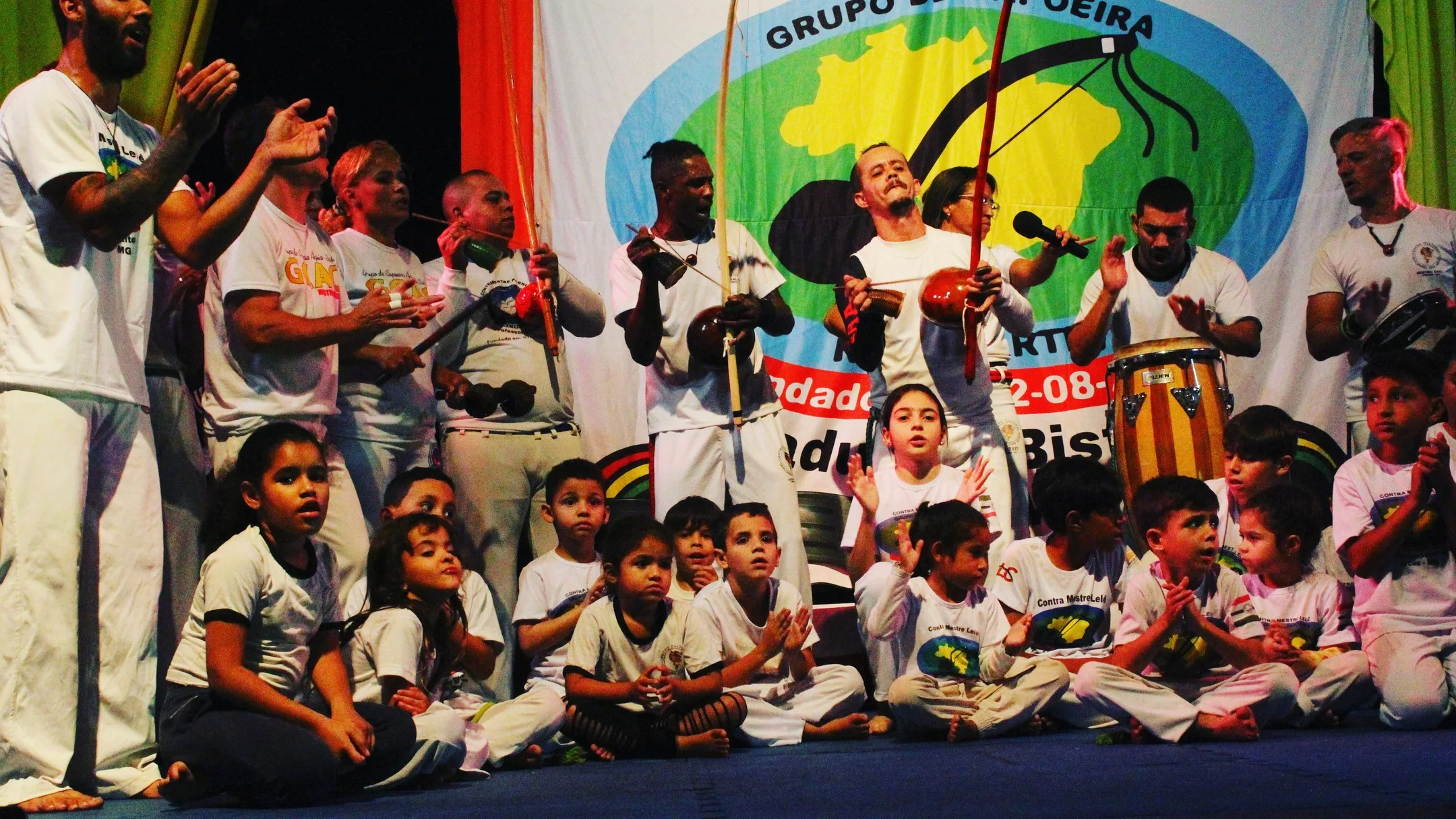 Roda de capoeira em um palco: adultos tocam berimbau, atabaque e pandeiro, e crianças de camiseta branca ficam sentadas à frente.