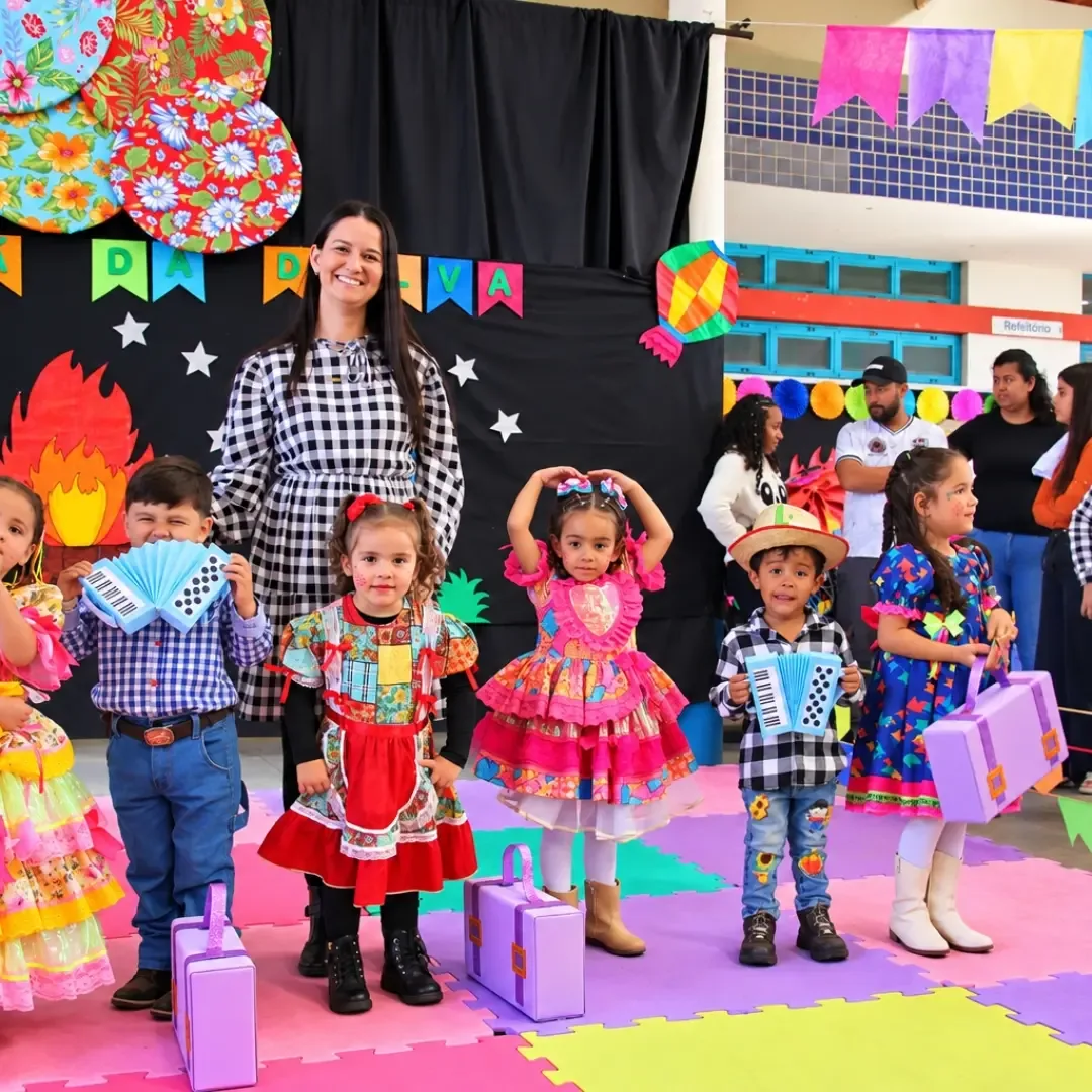 Crianças fantasiadas para uma festa junina posam em um ginásio, com uma mulher de vestido xadrez ao fundo.