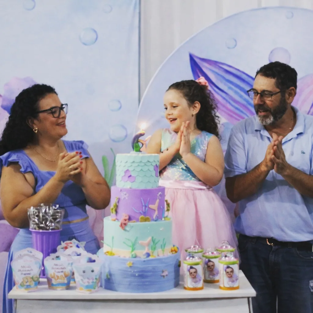 Menina bate palmas diante de um bolo de aniversário com uma vela acesa, ao lado de uma mulher e de um homem que também aplaudem.